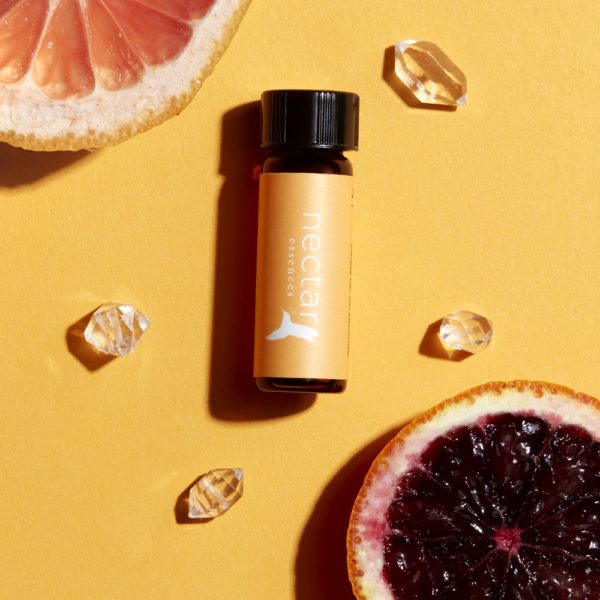 essential oil bottle with a bright orange 'nectar essences' label, centered between a ruby red grapefruit half, a dark red blood orange half, and four small clear citrine crystals, all on a sunny yellow background