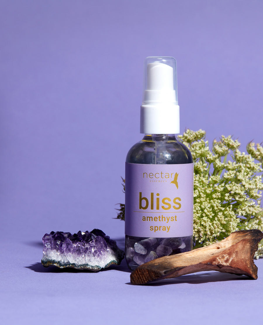 A spray bottle labeled 'bliss amethyst spray+' from Nectar Essences, accompanied by an amethyst crystal and a piece of wood, against a purple background.