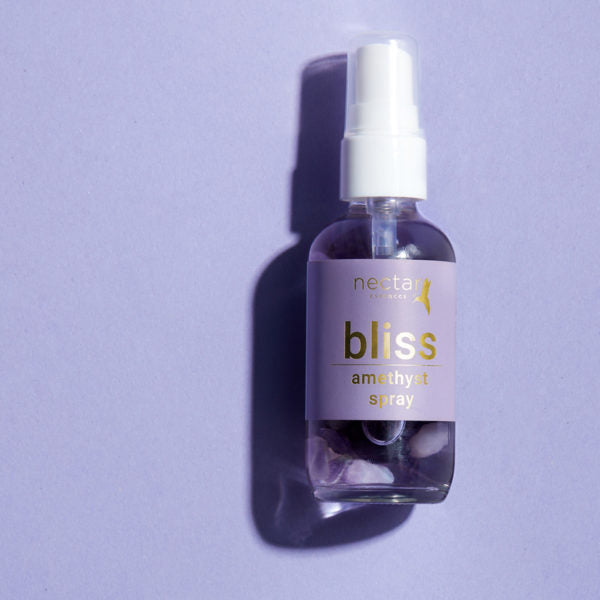 Close-up of a small, clear glass spray bottle with a white pump, labeled 'nectar essentials bliss amethyst spray.' The bottle is filled with a lavender-tinted liquid and contains small tumbled amethyst crystals visible at the bottom, set against a solid purple background.
