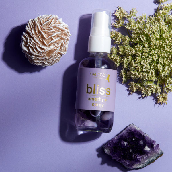 A spray bottle labeled 'bliss amethyst spray+' from Nectar Essences, accompanied by an amethyst crystal and a piece of wood, against a purple background.