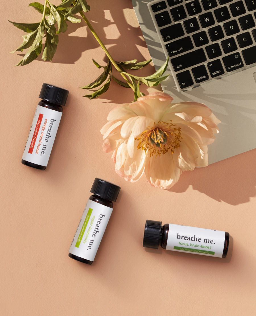 Three small bottles of genius kit essential oil with labels 'breathe me' arranged next to a laptop and a gold rose flower on a desk.
