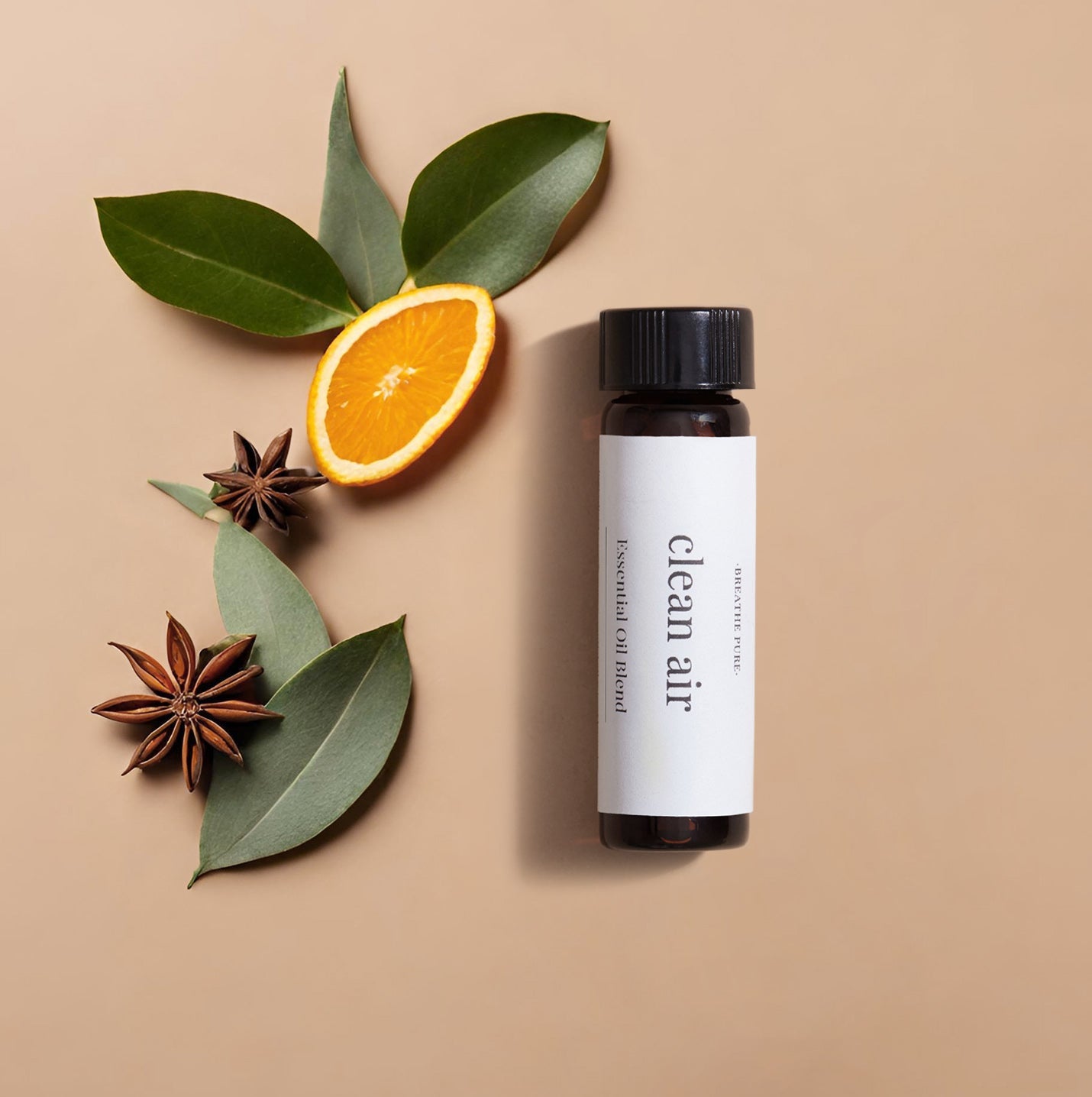 essential oil bottle with a white 'clean air' label, positioned next to eucalyptus leaves, star anise, and an orange slice on a tan background.