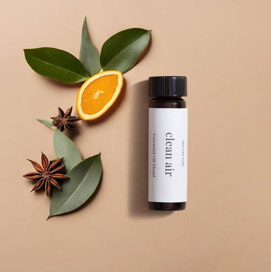 essential oil bottle with a white 'clean air' label, positioned next to eucalyptus leaves, star anise, and an orange slice on a tan background.