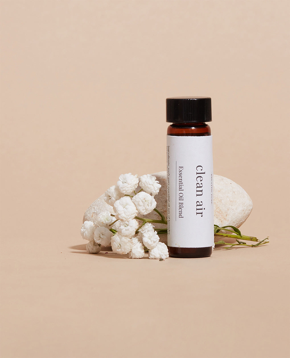 Close-up of the 'clean air' oil bottle on a white rock, with a small cluster of white baby's breath flowers on a light beige background.