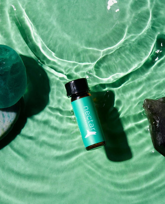 A small bottle of essential oil labeled 'nectar' floating on green water, surrounded by a splash of water and a few leaves.