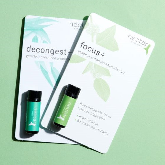 Two white product cards leaning on each other against a light green background. The front card displays the 'focus +' blend, with a small bottle of green-labeled essential oil. The text lists benefits: 'Pure essential oils, flower essences & lapis elixir,' 'Improves focus,' and 'Boosts memory & clarity.' The card underneath shows the 'decongest +' blend, with a teal-labeled oil bottle visible