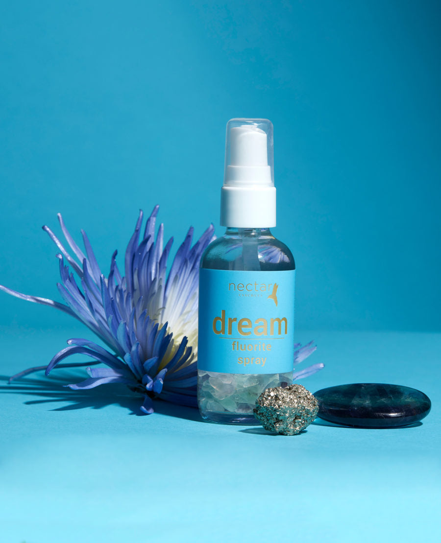 A clear spray bottle labeled 'dream' with a blue label and white spray nozzle, accompanied by a purple flower and a crystal on a blue background.