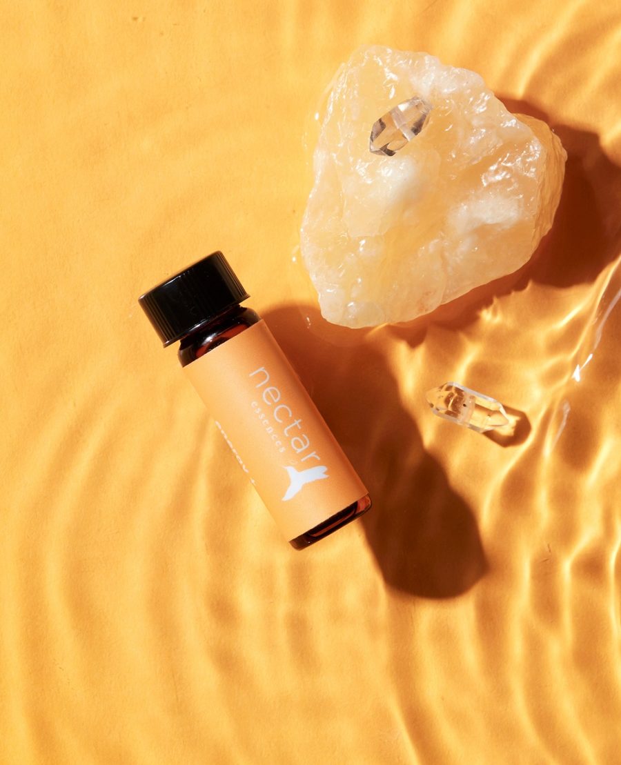 A small bottle of essential oil labeled 'nectar' is displayed next to a clear crystal on a surface with rippling light, suggesting a theme of aromatherapy and holistic wellness.