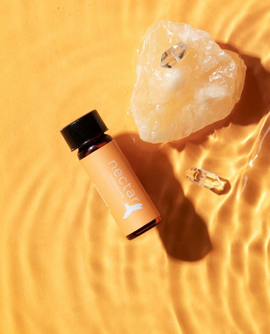A small bottle of essential oil labeled 'nectar' is displayed next to a clear crystal on a surface with rippling light, suggesting a theme of aromatherapy and holistic wellness.