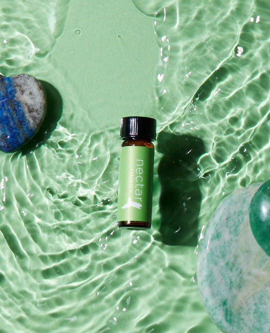 A small bottle of essential oil labeled 'focus+' placed on a water surface with ripples, alongside a blue stone and a green gemstone.