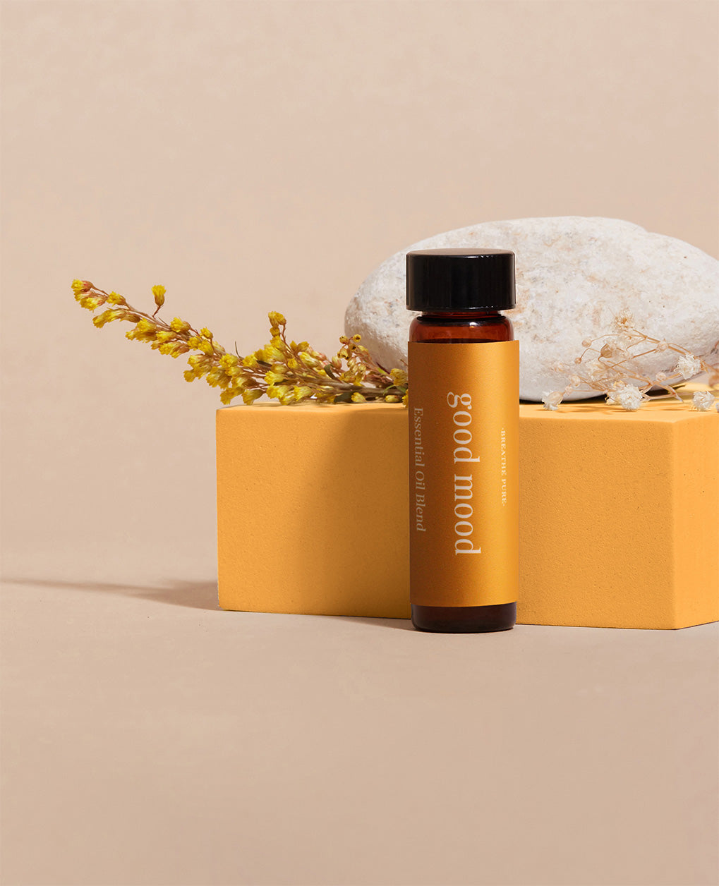 essential oil bottle with an orange 'good mood' label, set on an orange block next to yellow flowers and a white natural rock on a beige background.