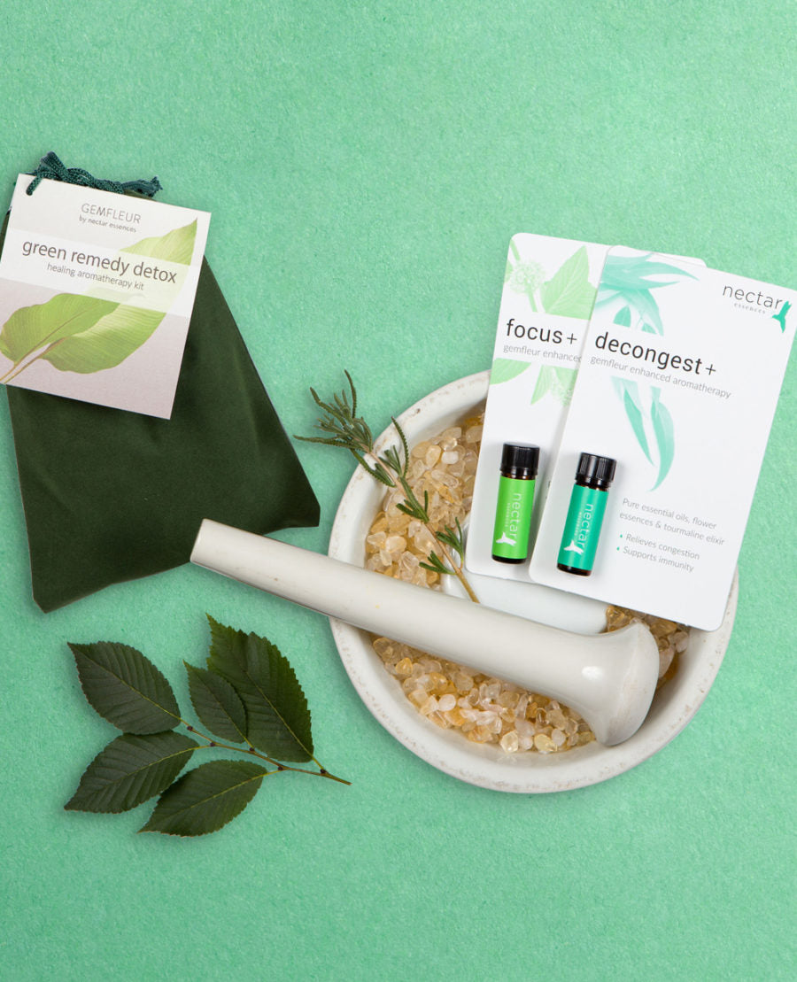 A collection of green-colored essential oil bottles and a detox pouch, displayed with a backdrop of green leaves and a white ceramic bowl.