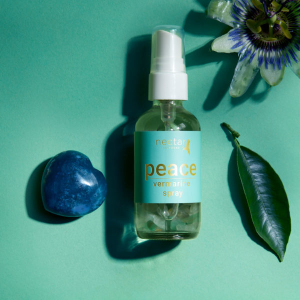 Clear spray bottle of 'nectar essentials peace vermarine spray' with a mint green label, containing green crystals, set on a teal background next to a polished blue heart-shaped stone, a green leaf, and a white and purple passion flower.