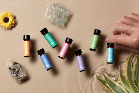 seven small 'nectar' essential oil bottles with labels in different colors (orange, teal, pink, light blue, purple, light green, dark green) scattered on a wet beige surface. The bottles are surrounded by small crystals, a yellow flower, and green foliage, with a hand reaching for one bottle