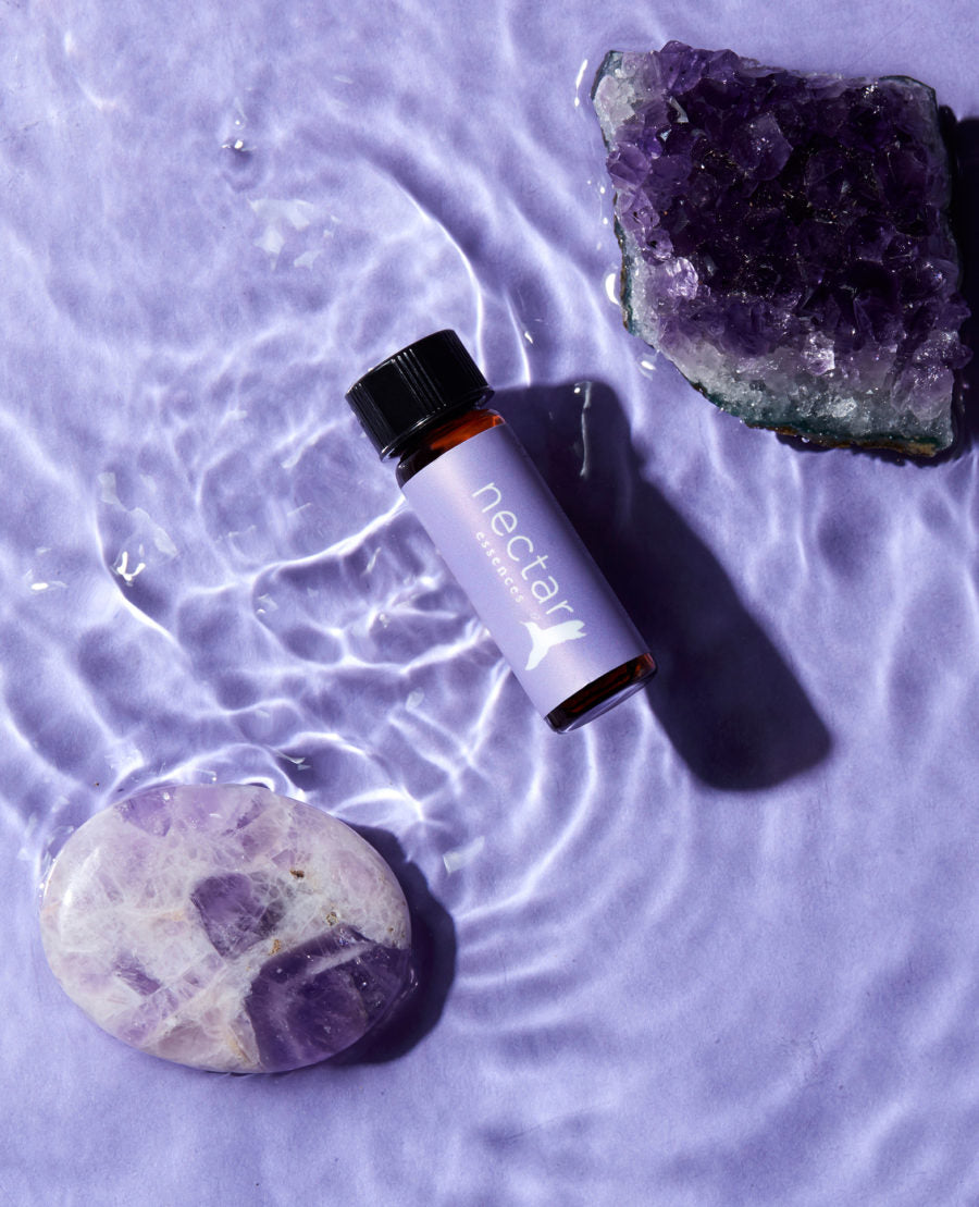 A bottle of essential oil lying on a surface with water ripples around, accompanied by an amethyst crystal and a piece of petrified wood.
