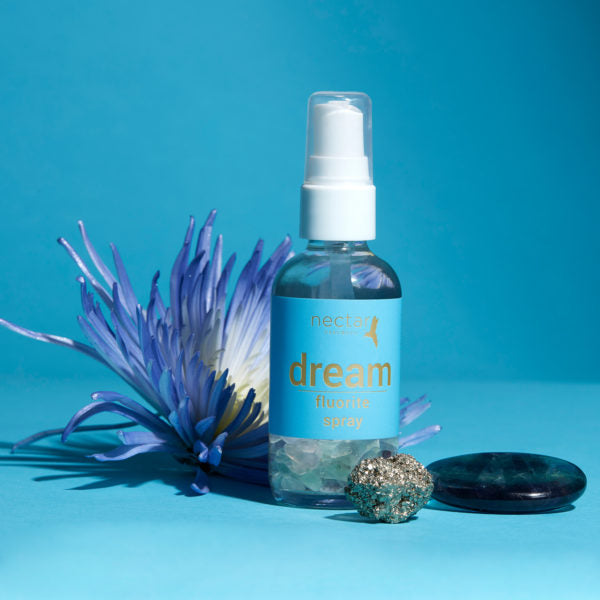 Clear spray bottle of 'nectar essentials dream fluorite spray' with a blue label, containing fluorite crystals, set on a blue background next to a large blue flower, a metallic pyrite stone, and a dark polished gemstone