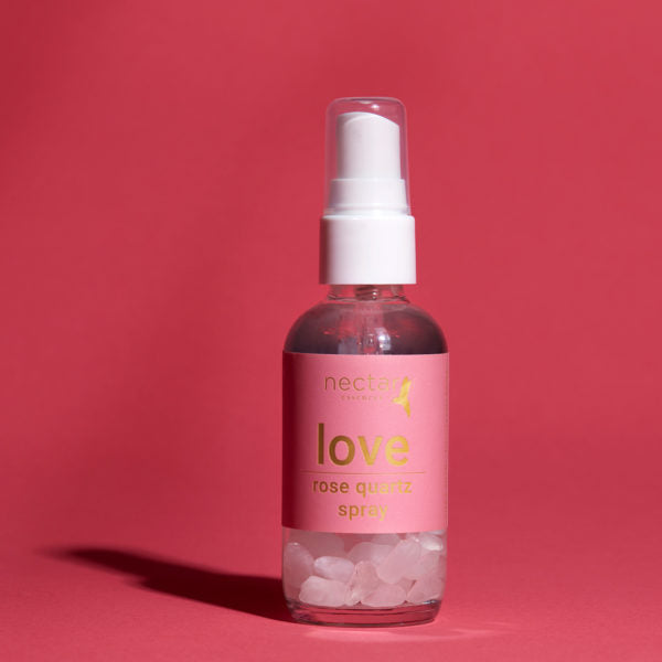 Vertical close-up of the small amber essential oil bottle against a vibrant pink background. The label is clearly visible, showing the white text 'love rose quartz spray' oriented vertically.