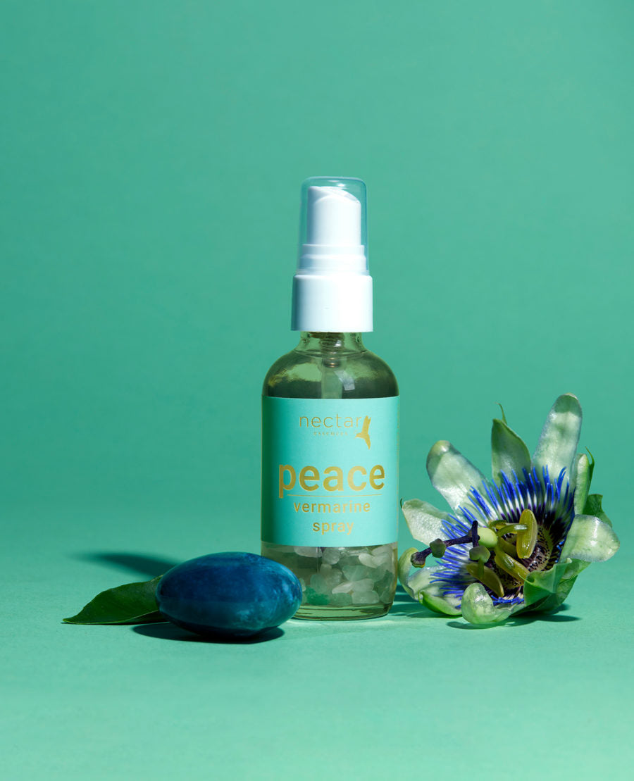 A clear glass bottle with a white spray nozzle labeled 'PEACE', accompanied by a blue barrel cactus flower and a purple passionflower on a green background.