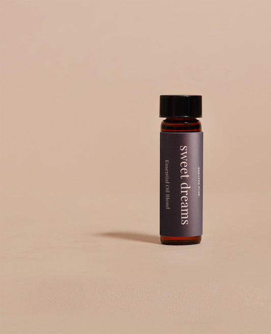 Close-up of Sweet Dreams essential oil blend glass bottle with a dark purple label on a simple beige background.