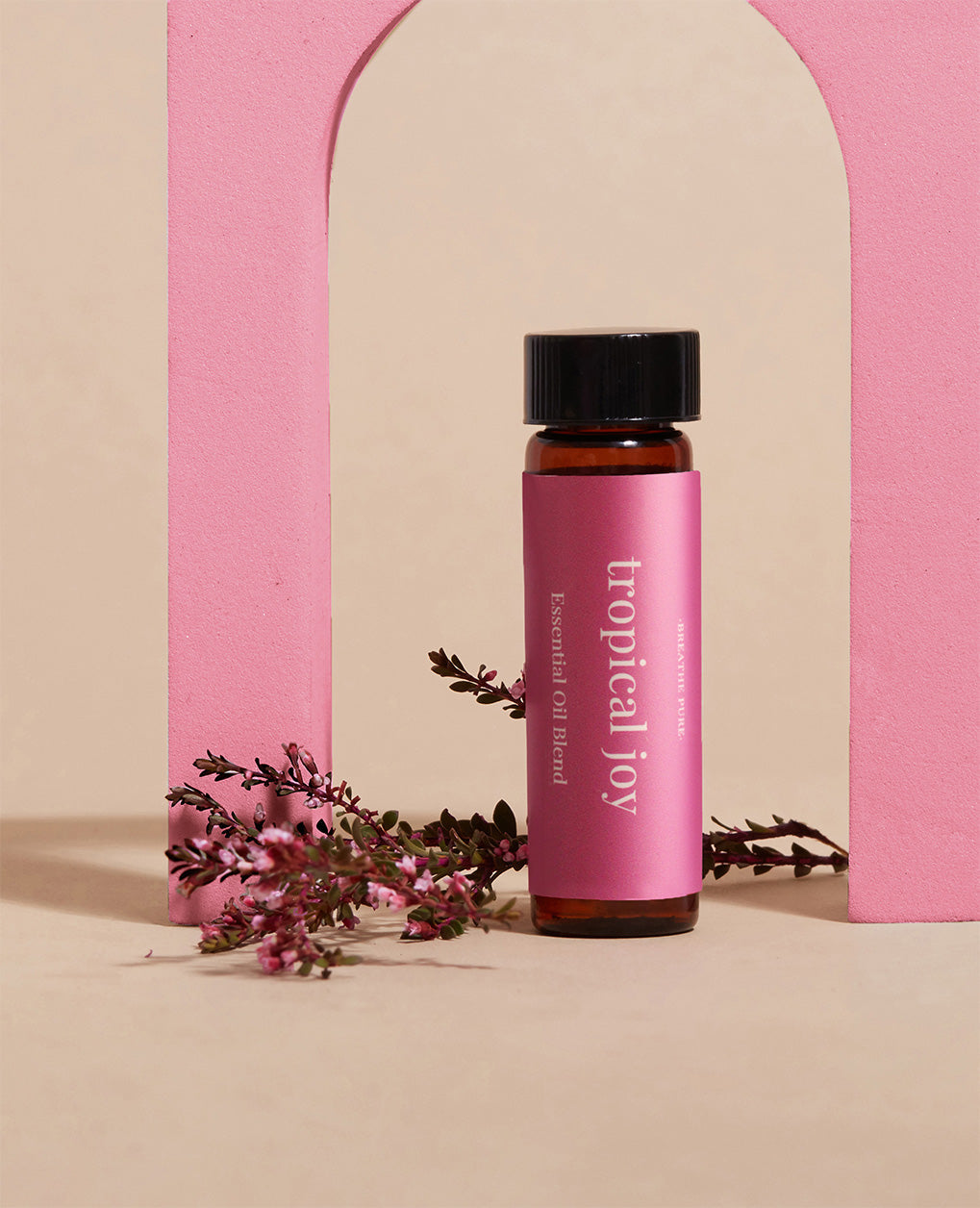essential oil bottle with a magenta 'tropical joy' label, framed by a pink foam arch with a sprig of pink flowers, on a neutral background