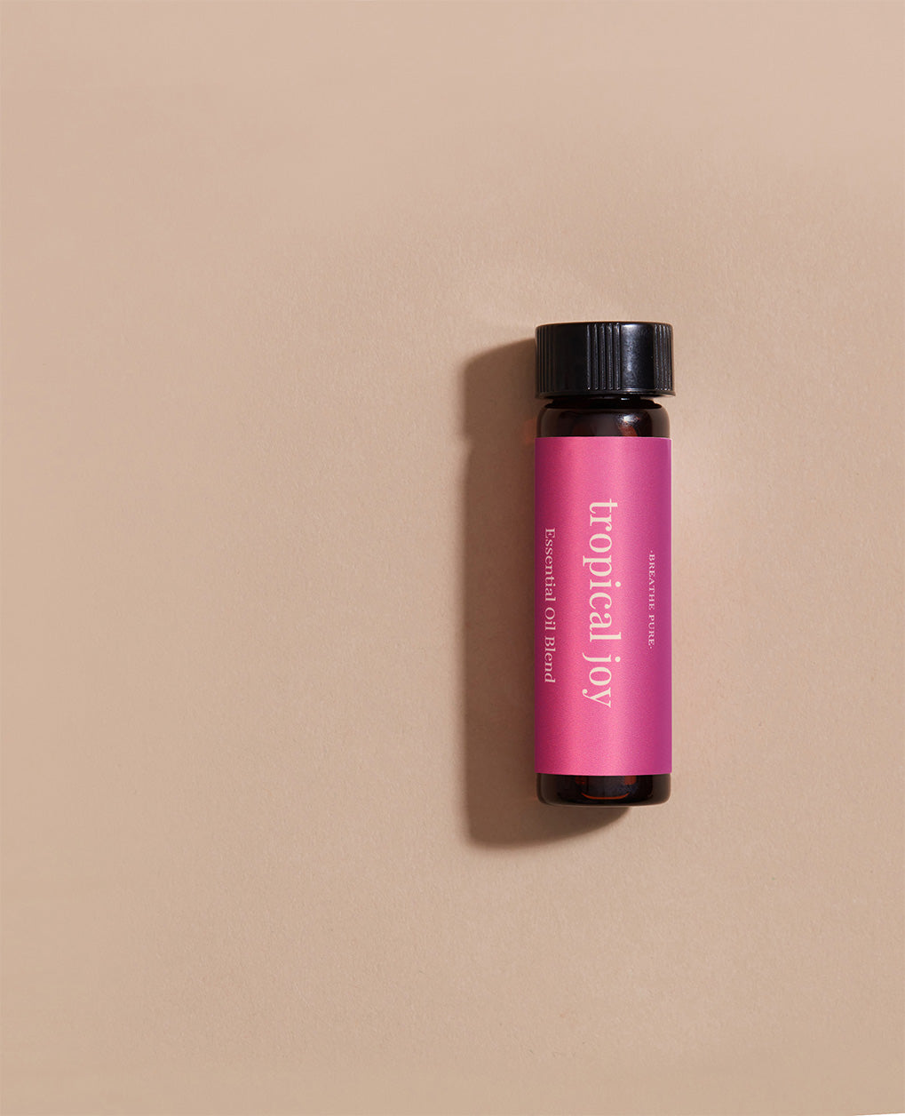 Vertical close-up of the small amber essential oil bottle laying gainst light pink background. The label is clearly visible, showing the white text 'tropical joy' oriented vertically.