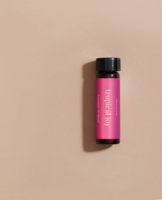 Vertical close-up of the small amber essential oil bottle laying gainst light pink background. The label is clearly visible, showing the white text 'tropical joy' oriented vertically.