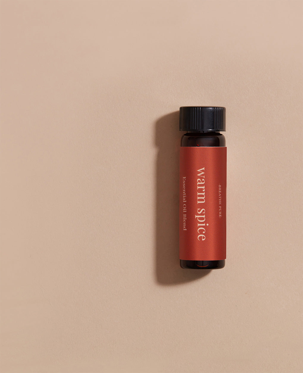 Vertical close-up of the small amber essential oil bottle laying gainst light pink background. The label is clearly visible, showing the white text 'warm spice' oriented vertically.