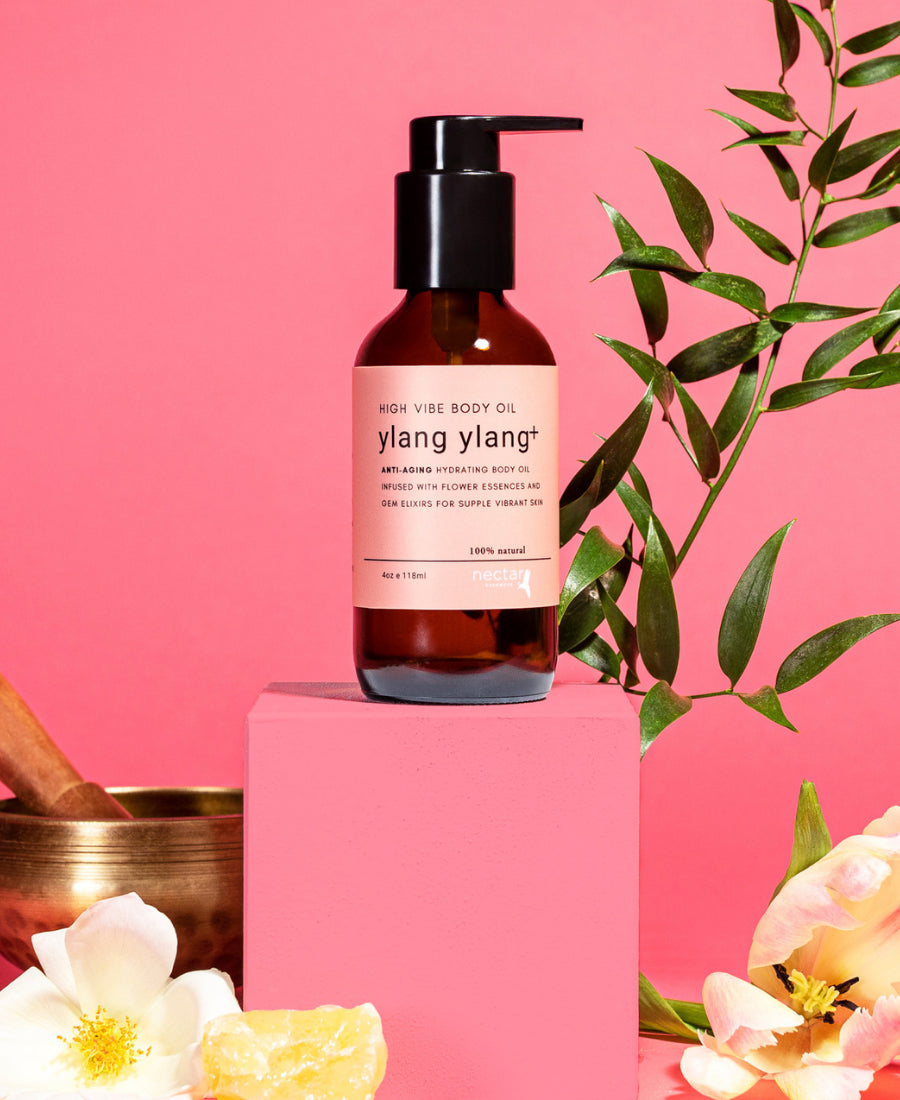 A bottle of Ylang Ylang+ High Vibe Body Oil displayed on a pink pedestal with floral and botanical decorations in the background.
