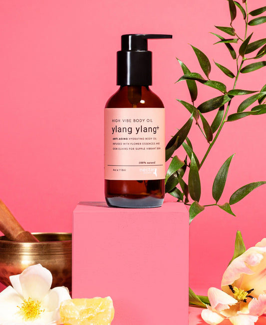 A bottle of Ylang Ylang+ High Vibe Body Oil displayed on a pink pedestal with floral and botanical decorations in the background.