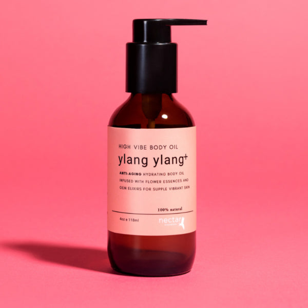 High Vibe Ylang Ylang Plus Anti-Aging Hydrating Body Oil in an amber glass pump bottle on a vibrant pink background