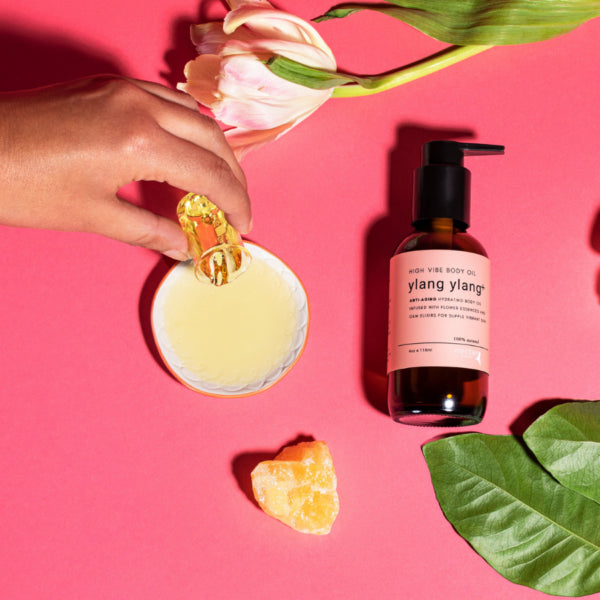 Top-down view of Ylang Ylang Body Oil being poured into a dish next to an orange crystal and a flower, demonstrating application.