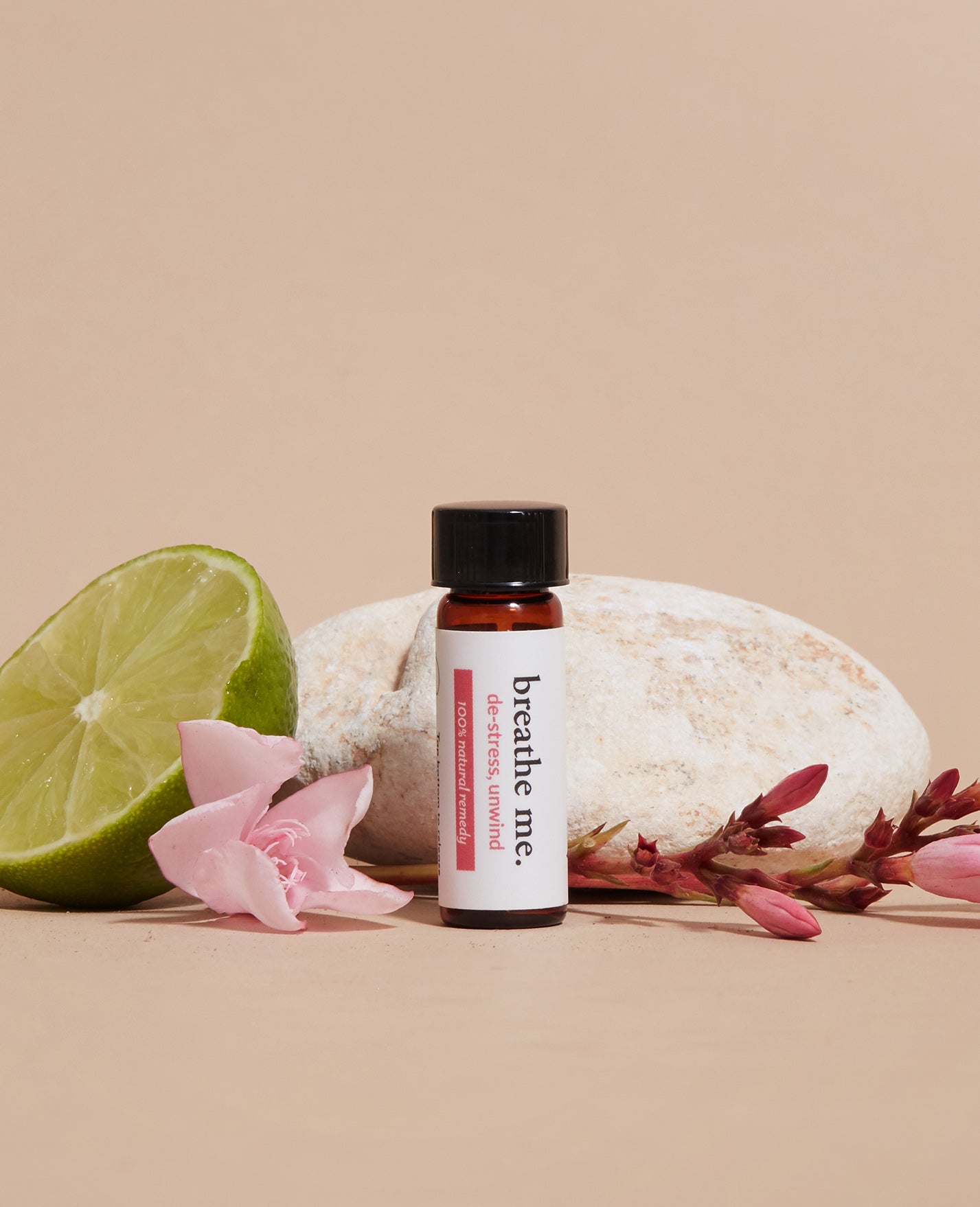 A travel-sized bottle labeled 'Breathe Me' with a brown cap, placed on a surface with a green lime and pink flower petals around it.