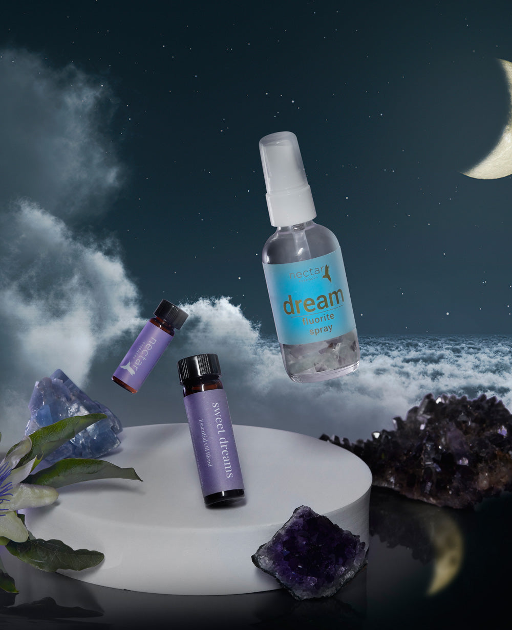 A aromatherapy kit including a spray bottle labeled 'dream', two small bottles of essential oils, and surrounding by what appears to be lavender plants and crystals, presented against a night sky backdrop.